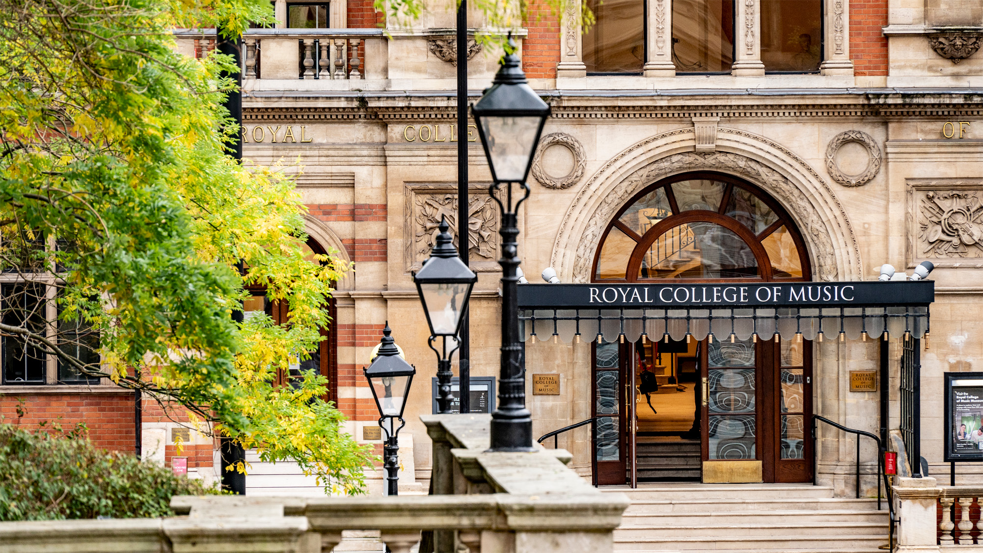 The entrance of the Royal College of Music, framed by black lampposts and greenery on the left, with an open door.